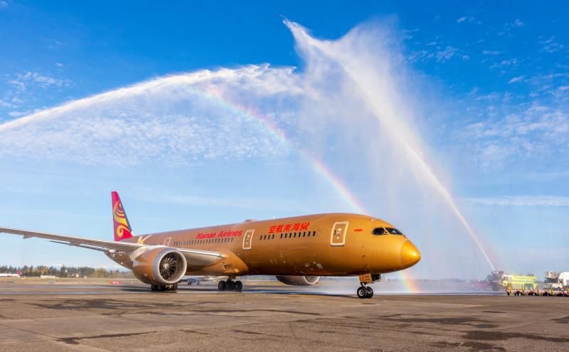A gold-colored commercial airplane on an airport runway passes beneath crossing arcs of water sprayed by fire trucks, creating a visible rainbow against a bright blue sky.