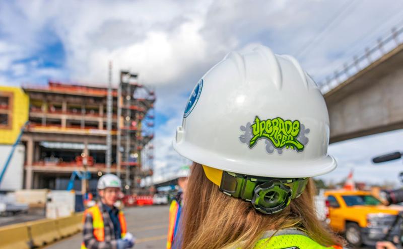 Worker with a construction hat that has the Upgrade SEA logo while a construction project is in progress in the background. 