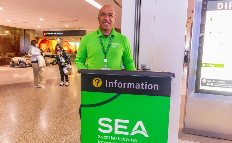 Information desk volunteer in a bright green SEA Seattle‑Tacoma International Airport shirt stands behind an information podium in the terminal, with travelers and signage in the background.