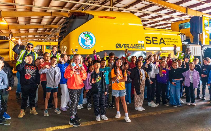 Large group of children and airport staff standing and waving in front of a yellow Seattle‑Tacoma International Airport snow removal vehicle inside a maintenance hangar.