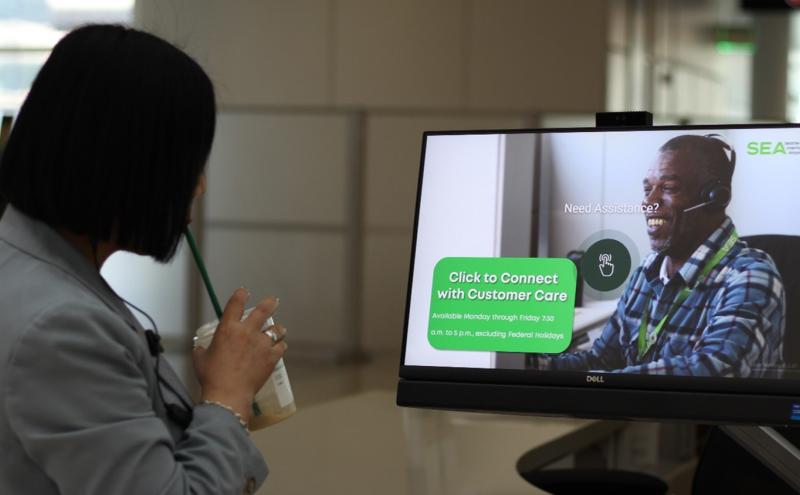 Traveler standing in front of a touchscreen kiosk displaying a SEA Airport customer‑care screen with a representative shown on the monitor.