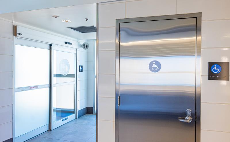 An accessible restroom entrance with a metal door displaying an accessibility symbol, next to frosted sliding doors leading to a restroom area.