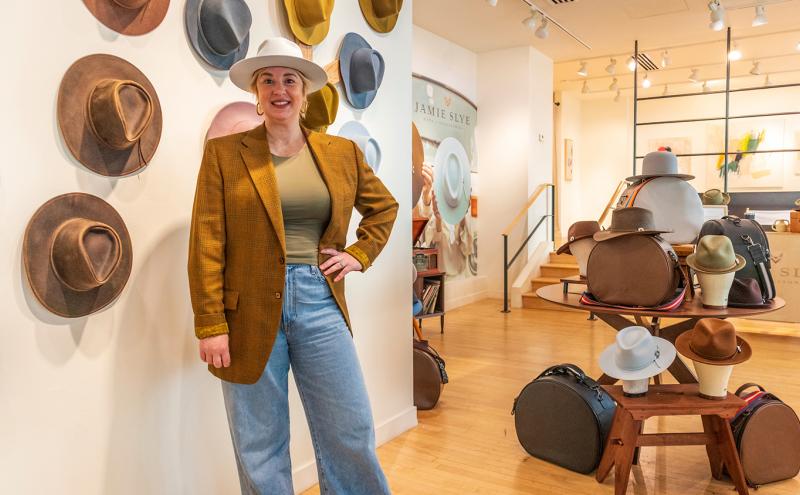 Jamie Slye stands beside a wall display of hats in a bright hat shop, with more hats and hat cases arranged on tables in the background.