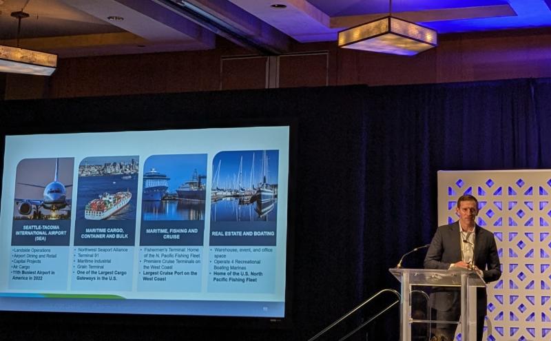 Commissioner Calkins stands at a podium in a conference room, presenting beside a large projection screen. The screen displays four labeled sections with images representing Seattle‑Tacoma International Airport, maritime cargo and container operations, maritime fishing and cruise, and real estate and boating. Hanging light fixtures and a decorative backdrop are visible in the room.