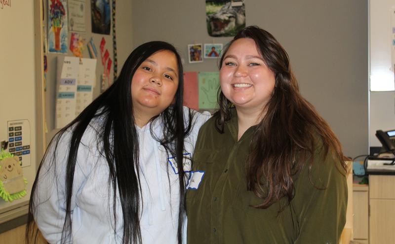 Two people standing side by side in a classroom, with educational materials and a whiteboard visible in the background.