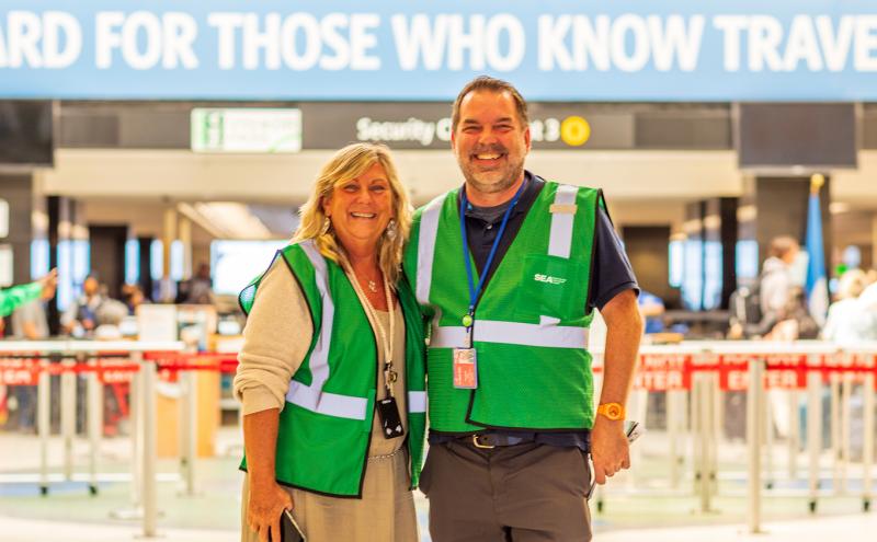 Two people wearing green safety vests stand together in an airport security area.