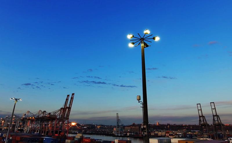 A tall pole with bright lights stands over a shipping port with cranes and containers at dusk.