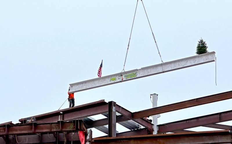 A steel beam with an American flag and a small evergreen tree is lifted by a crane onto a building frame, while a worker stands on the structure guiding it.