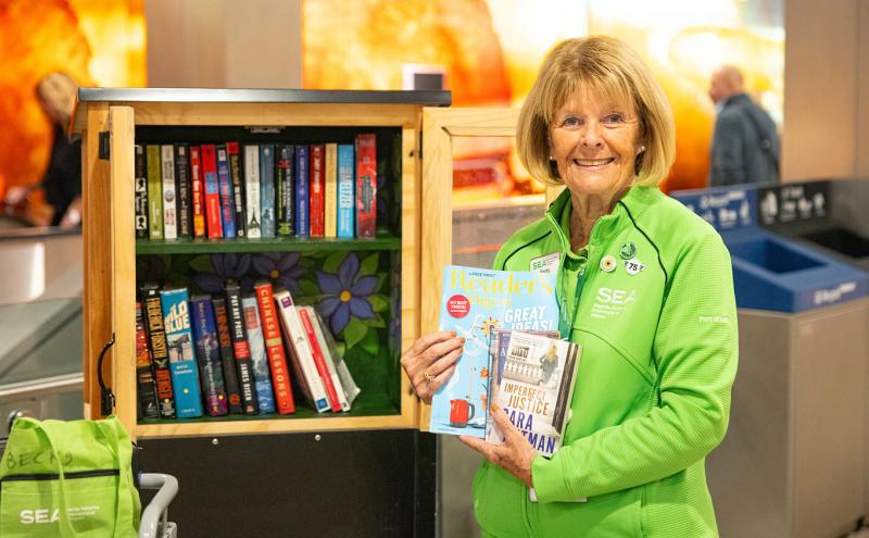 Becky Nickels stands beside a small wooden book-sharing cabinet and holds a few books and magazines.