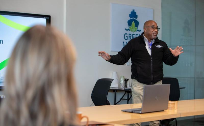 Delmas Whittaker stands at the front of a meeting room giving a presentation, with a laptop on the table and a screen displaying slides in the background.