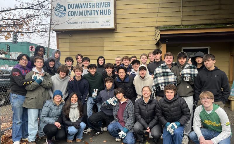 A large group of people pose together outside a building with a sign that reads “Duwamish River Community Hub.” Trees, a fence, and a street intersection are visible in the background.