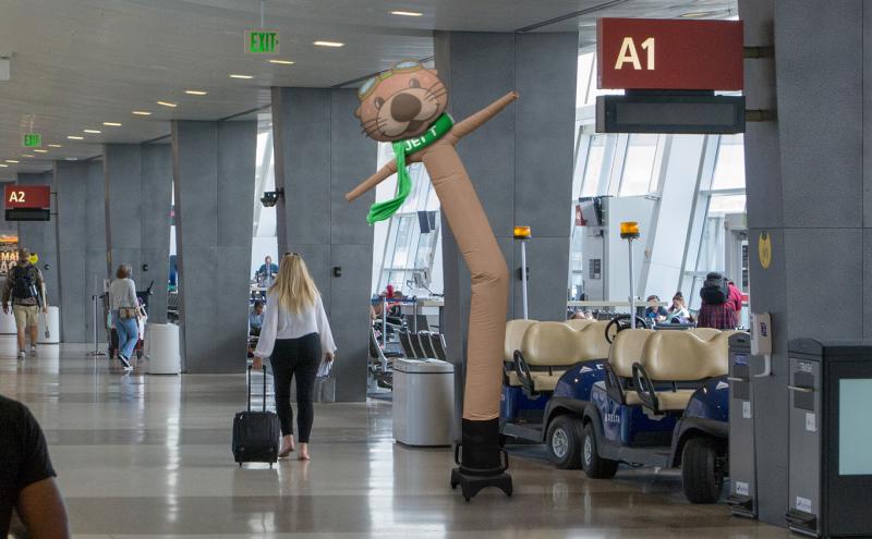 An airport concourse with gate signs and travelers, featuring a tall inflatable otter‑themed tube figure near seating and carts.
