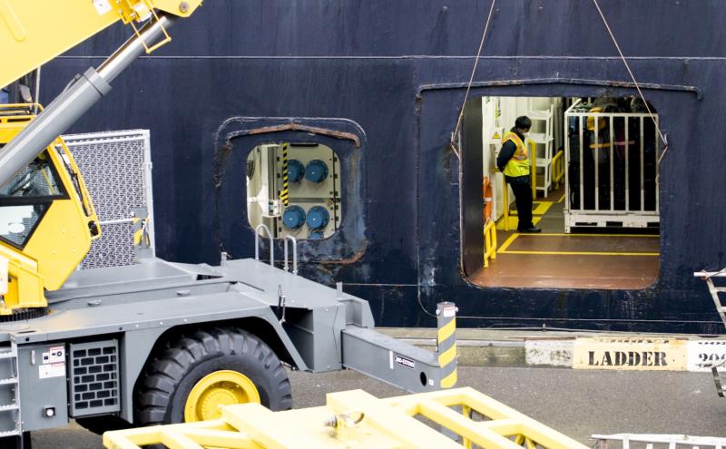 Cargo operation scene showing a large yellow crane positioned beside an open doorway on a ship, with a worker in a safety vest standing inside the vessel.