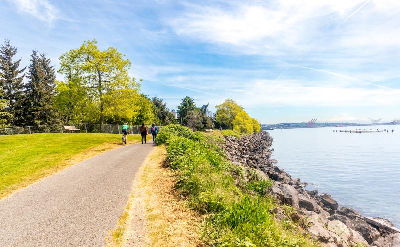 Pathway at Centennial Park beside rocky shoreline and calm water, with people walking and cycling and trees along the edge.