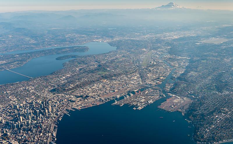 High-altitude aerial view of a coastal city with lakes, waterways, and a distant snow-capped mountain.