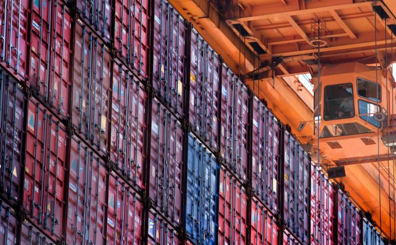 Rows of stacked red shipping containers viewed from below with an orange crane and operator cabin overheard