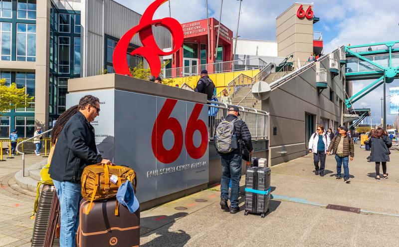 People with rolling luggage walk near a waterfront terminal building marked with a large red “66” sign.