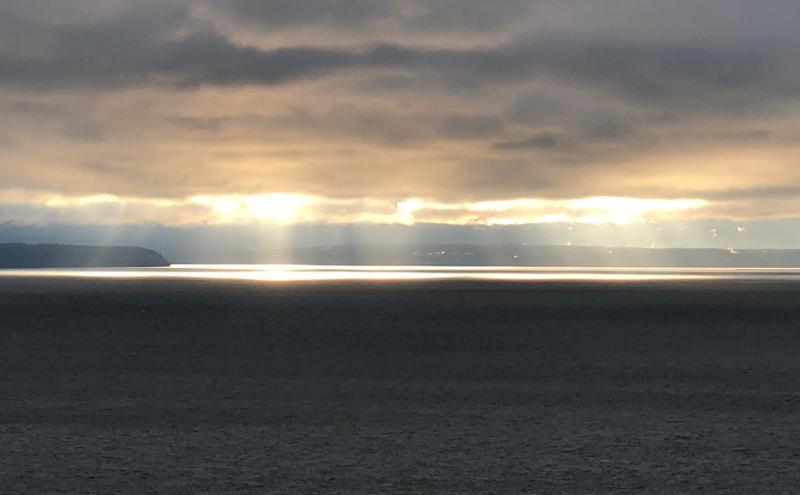 Sunlight breaks through heavy clouds over a wide expanse of water, creating bright reflective bands on the surface. A distant shoreline sits low on the horizon beneath the glowing sky.
