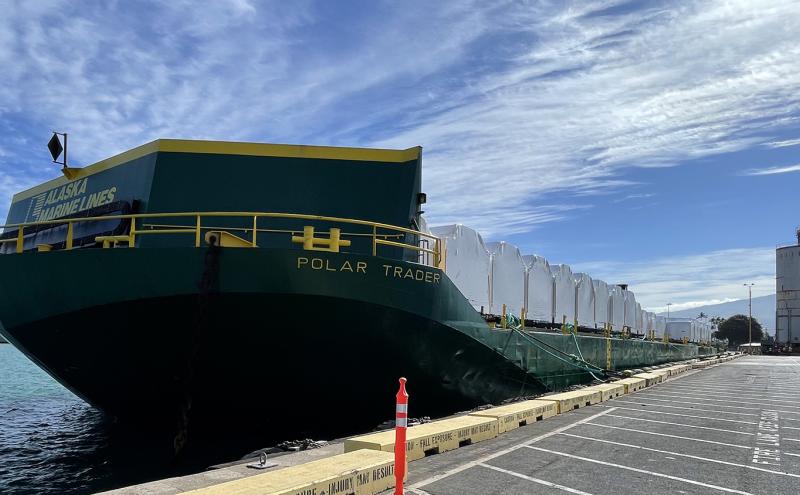 A cargo barge labeled “POLAR TRADER” is docked at a port with white-wrapped freight on deck under a partly cloudy sky.