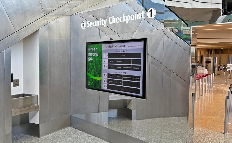 Airport security checkpoint with a digital sign showing lane status and queue stanchions in front.