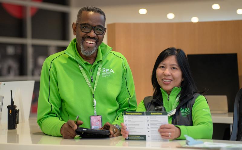 Two SEA Airport staff members stand behind an information counter, wearing matching bright green uniforms with SEA branding. One holds a device while the other holds a printed information brochure labeled ‘Transportation’ and ‘Amenities.’ A two‑way radio and other desk items are visible on the counter, with a softly lit interior background behind them.