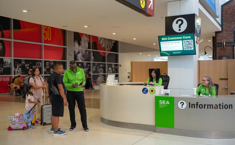 An airport information desk area where SEA Customer Care staff assist travelers. A staff member in a bright green SEA uniform speaks with a group of travelers who have luggage, including a small colorful rolling suitcase. Two additional staff members sit behind the curved information counter, which displays question‑mark signage and SEA branding. A digital screen above the desk provides SEA Customer Care details and a QR code. The background features a red and gray wall with photos and graphics, along with 