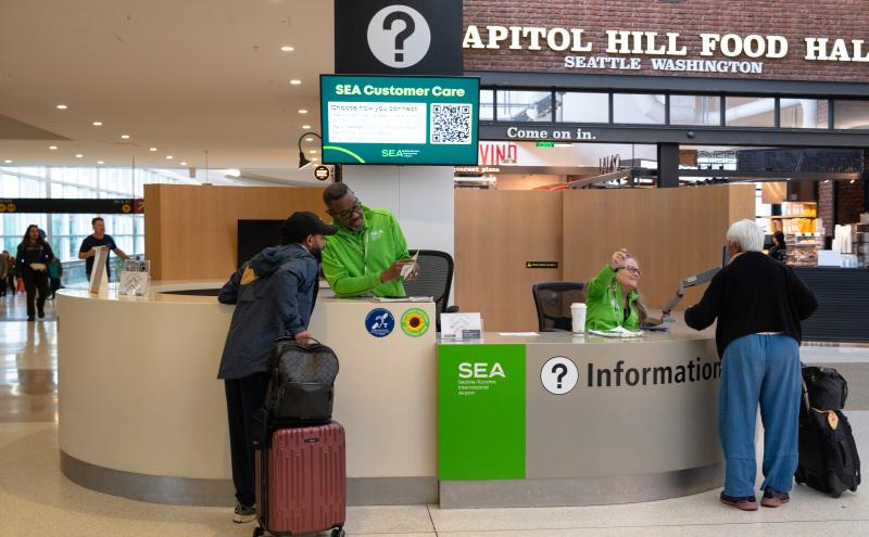 An airport information desk with SEA Customer Care staff assisting travelers. One staff member in a green SEA uniform speaks with a traveler who has a suitcase and backpack. Another staff member behind the counter gestures toward a traveler using a mobility aid. A large question‑mark sign and digital display with SEA Customer Care information and a QR code hang above the desk. In the background is the Capitol Hill Food Hall with visible signage and storefronts.