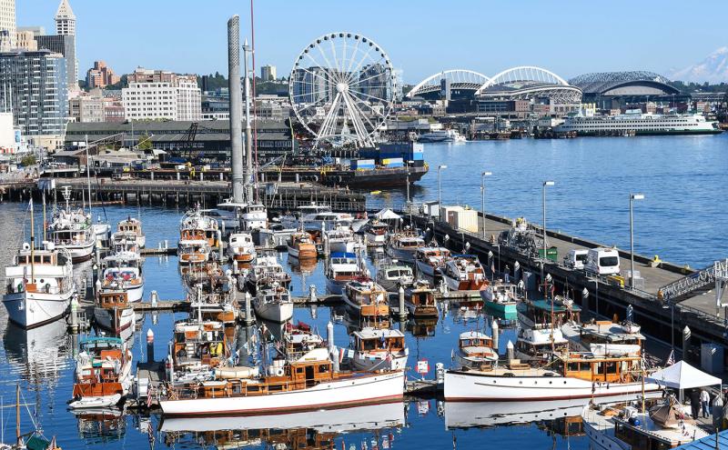 A marina filled with numerous boats docked along the piers, with a waterfront cityscape in the background that includes tall buildings, a large Ferris wheel, stadium structures, and a distant snow-covered mountain across the water.