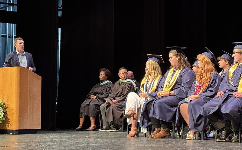 A speaker stands at a podium during a graduation ceremony, while a group of graduates in caps and gowns sit on stage beside several seated faculty or staff members.