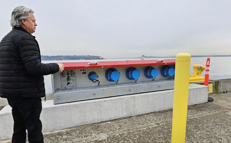 Person standing on a waterfront pier beside a large metal utility box with multiple blue connectors, with water and distant shoreline in the background.