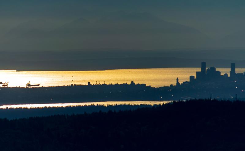 A distant city skyline appears in silhouette above a sunlit bay, with ships and cranes visible along the waterfront.