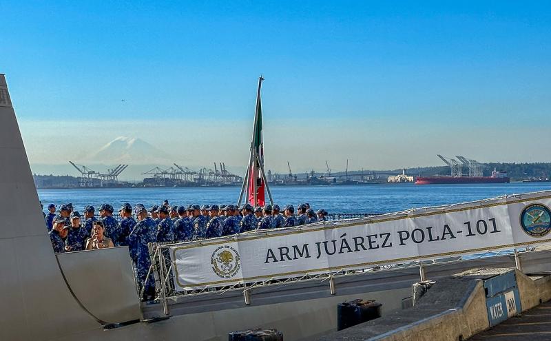 A naval vessel at a waterfront with uniformed crew members gathered on deck near a banner reading “ARM Juárez POLA-101,” with port cranes and a distant mountain in the background.