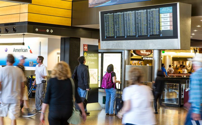 Busy airport concourse with travelers walking and checking a large overhead flight information display.