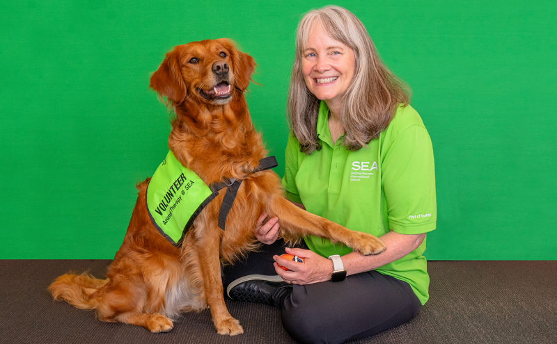 A person in a green SEA Airport shirt sits beside a golden dog wearing a “Volunteer Animal Therapy @ SEA” vest.