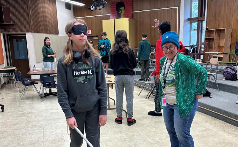 A classroom where a person wearing a blindfold uses a white cane while another person stands nearby, with several others and chairs in the background.