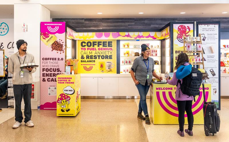 Customers and staff at a brightly colored coffee and gift kiosk in an airport, with signs promoting specialty coffee and products.