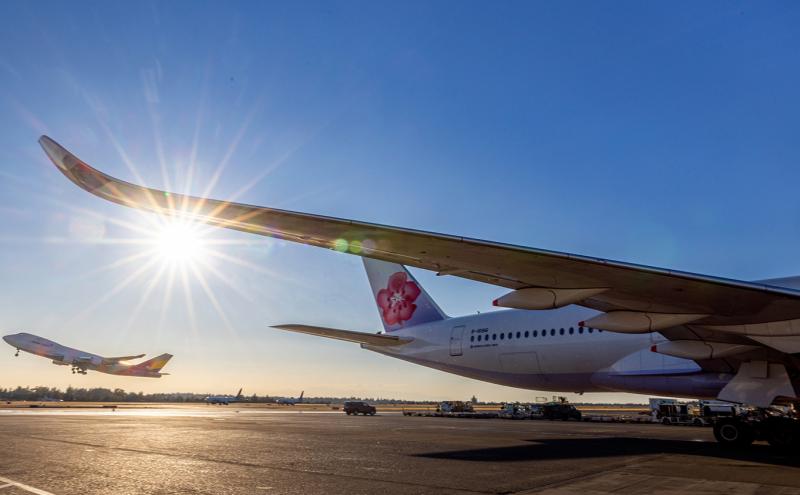 A large aircraft sits on the tarmac while another plane takes off in the distance under a bright sun.