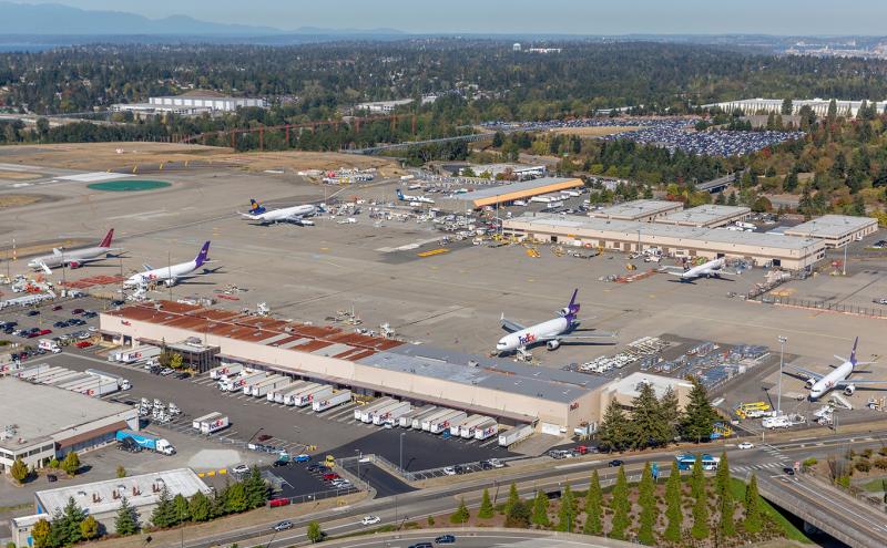 A wide aerial view of SEA Airport showing multiple cargo aircraft on the tarmac, airport service vehicles, and surrounding buildings and parking areas.