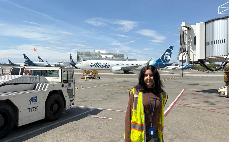 Isabella Newcomer wearing a reflective safety vest stands on an airport tarmac near a jet bridge, with several Alaska‑branded airplanes and ground service vehicles in the background.
