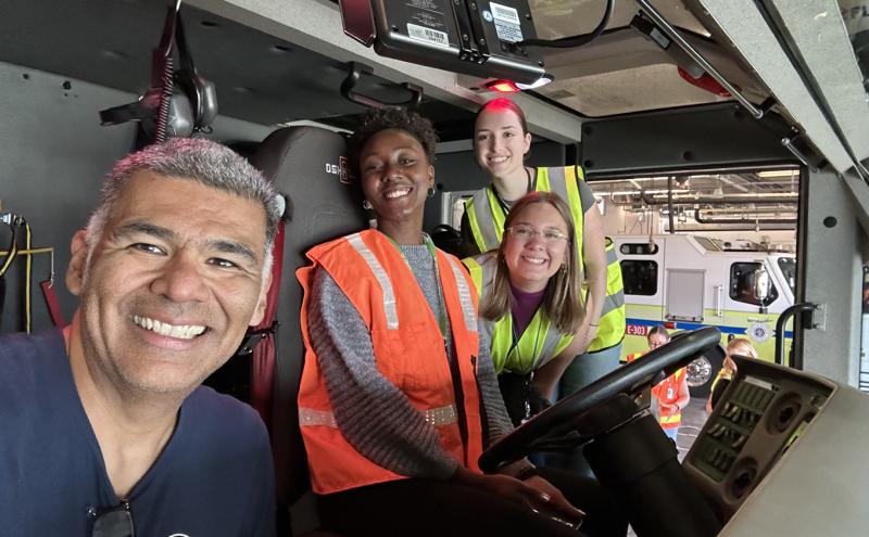 Four people wearing safety vests inside an emergency vehicle cab, with the dashboard and a fire truck visible in the background.