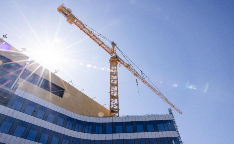 A tall yellow construction crane rises above a modern building with curved blue‑and‑white paneling. Bright sunlight shines behind the crane, creating a strong lens flare across the image. The sky is clear and blue.