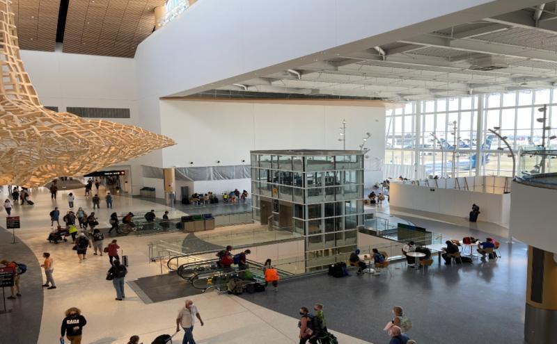 View from the N Concourse showing a large airport hall with travelers, escalators, tall windows facing aircraft, and a suspended wooden art structure.