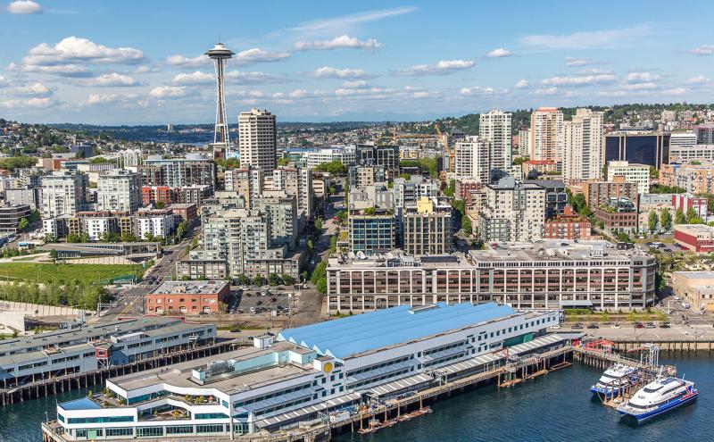 A waterfront cityscape with a long pier building, two docked vessels, and a dense skyline with a tall observation tower in the background.