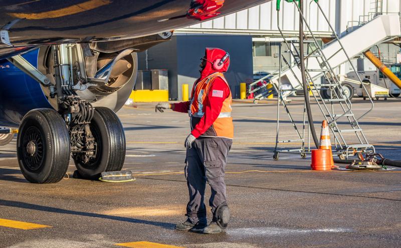An airport ground crew member wearing protective gear and a high‑visibility vest stands near the landing gear of a parked aircraft on the tarmac, inspecting the underside of the plane. Nearby are service equipment, a set of mobile stairs, and traffic cones.