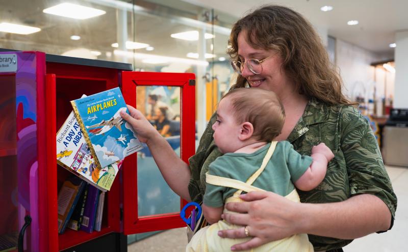 An adult holding a small child selects a children’s book from a red mini‑library box.