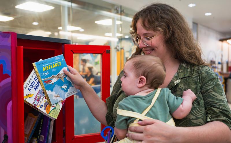 A person holding a young child selects a book from a small red library cabinet filled with children’s books inside an indoor airport area.