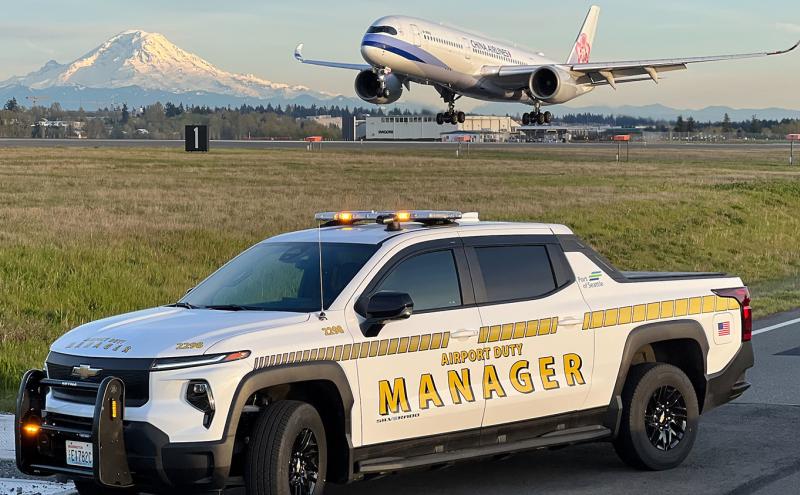 White pickup truck marked ‘Airport Duty Manager’ sits beside a runway with its roof lights on. In the background, an airplane is descending toward the runway, and a snow-covered mountain rises behind the landscape.