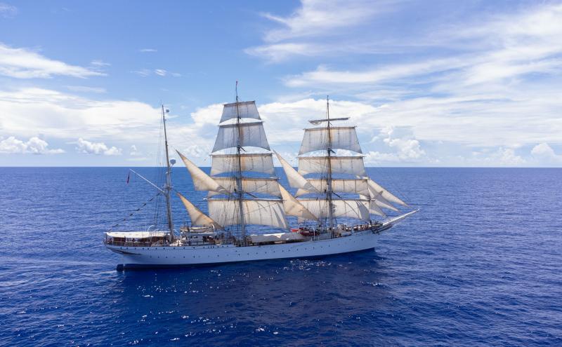 A large white sailing ship with multiple masts and cream-colored sails travels across a calm blue ocean under a bright sky with scattered clouds.
