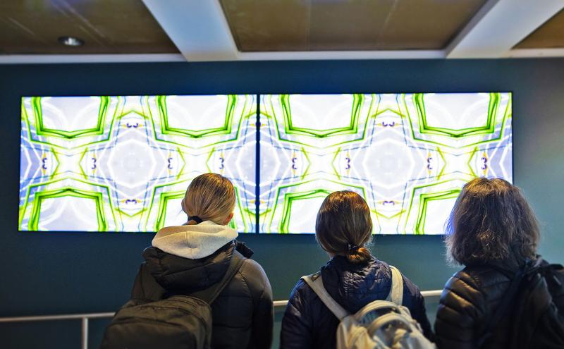 Three individuals with backpacks stand indoors facing two large, brightly lit digital screens displaying symmetrical green and white abstract patterns.