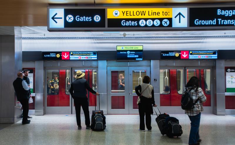 Passengers with rolling luggage stand on a polished floor facing closed doors of an airport underground shuttle train. Overhead signs display directions, including arrows toward Gates D and Gates A, B, S, and a yellow line indicator. The train’s interior and a person inside are visible through the glass doors.